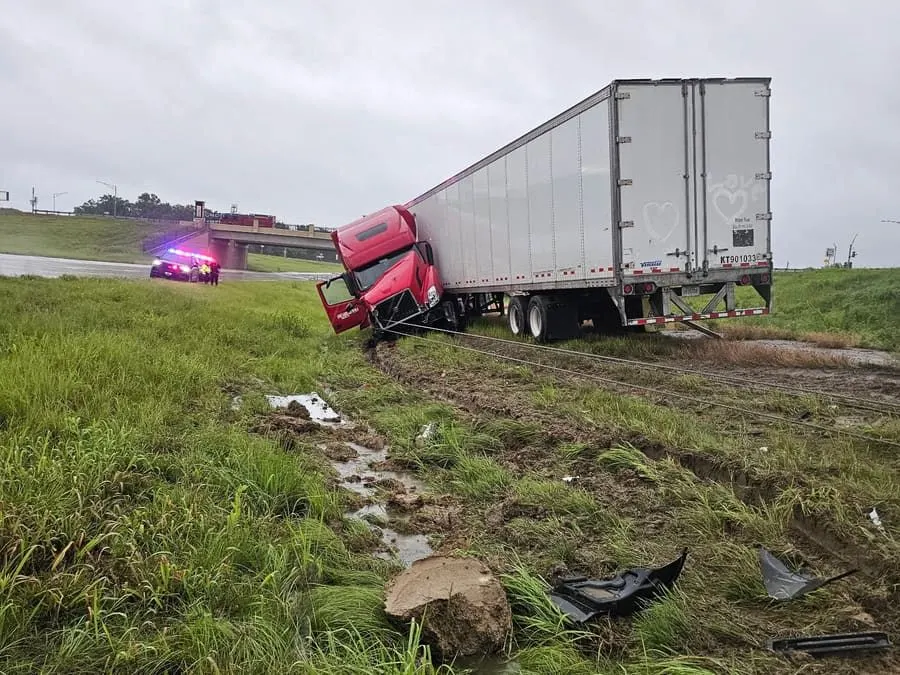 A red semi tractor and loaded dry van off the road in a wet ditch, recovery lines rigged to the tractor, with police on the shoulder above.